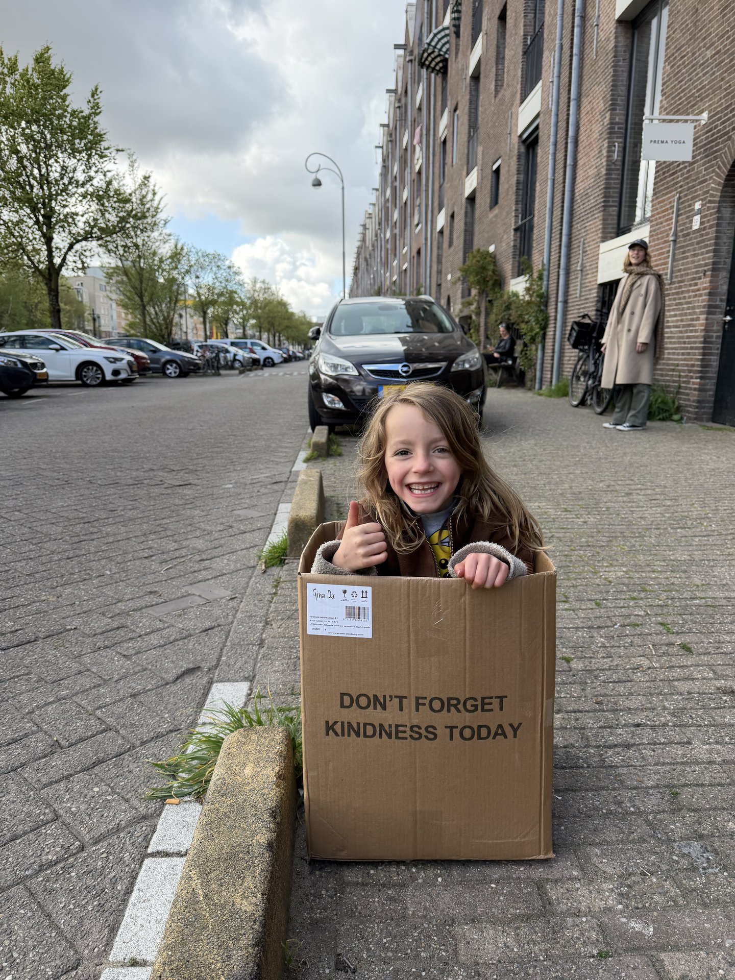 A joyful child sits inside a cardboard box, giving a thumbs-up on a street in Entrepotdok, Kadijken, Amsterdam